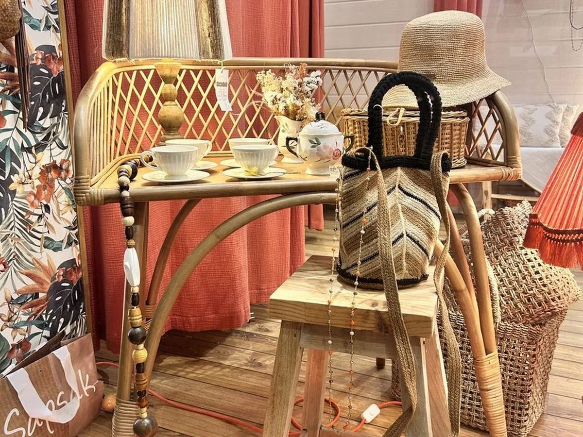 Coin salon en rotin : banquette, guéridon, chapeau et sac posés, rideau fleuri en fond.