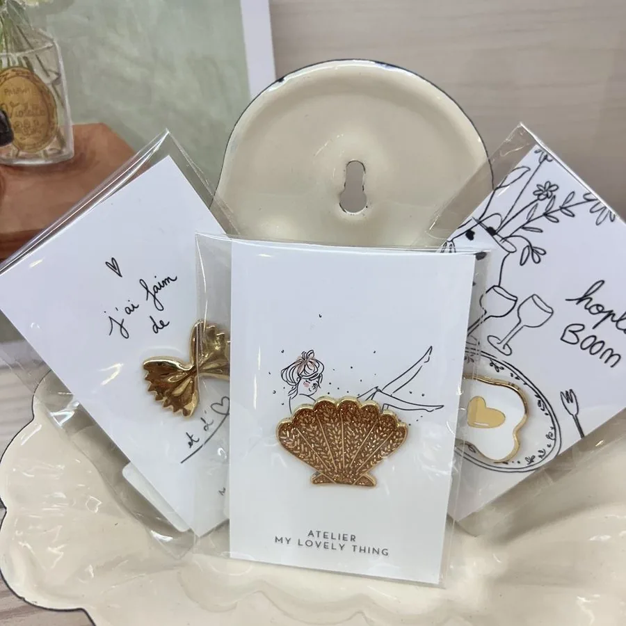 Carte brodée d’un coquillage doré, posée devant une céramique blanche.